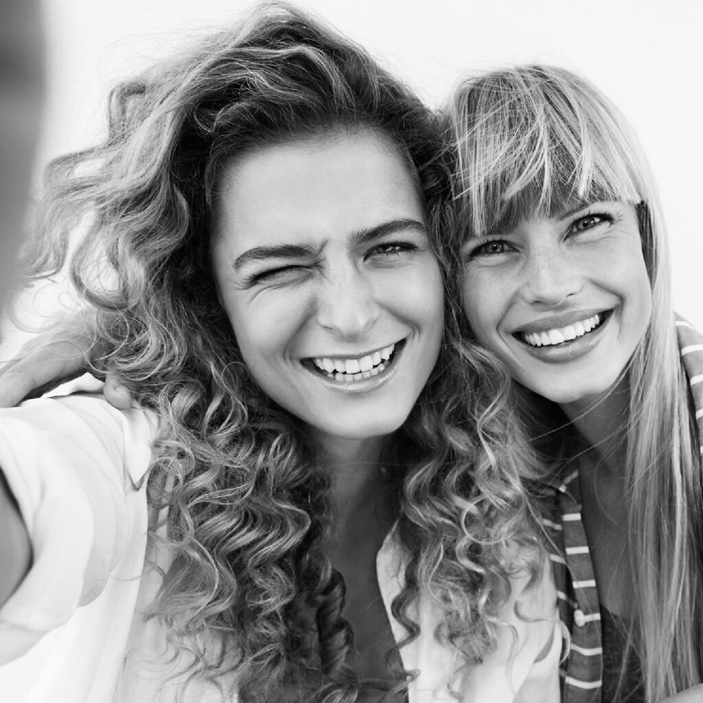 Zwei junge Damen mit gepflegten Haaren schauen fröhlich in die Kamera